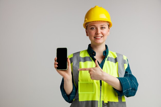 Sorridente giovane operaio edile femminile che indossa il casco di sicurezza e giubbotto di sicurezza che tiene e che indica al telefono cellulare