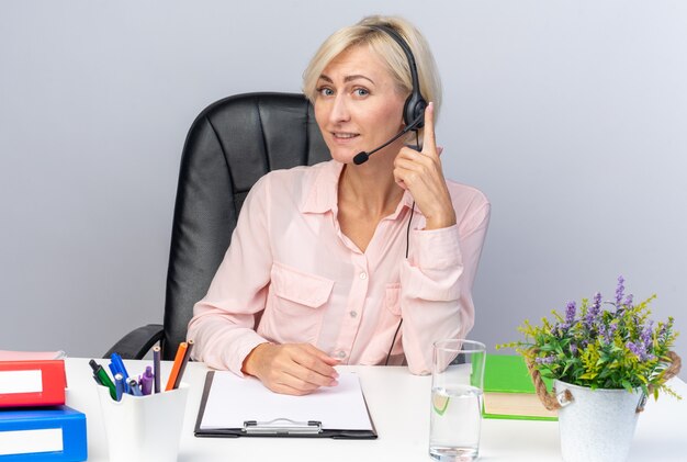 Sorridente giovane operatore di call center femminile che indossa l'auricolare seduto al tavolo con strumenti per ufficio