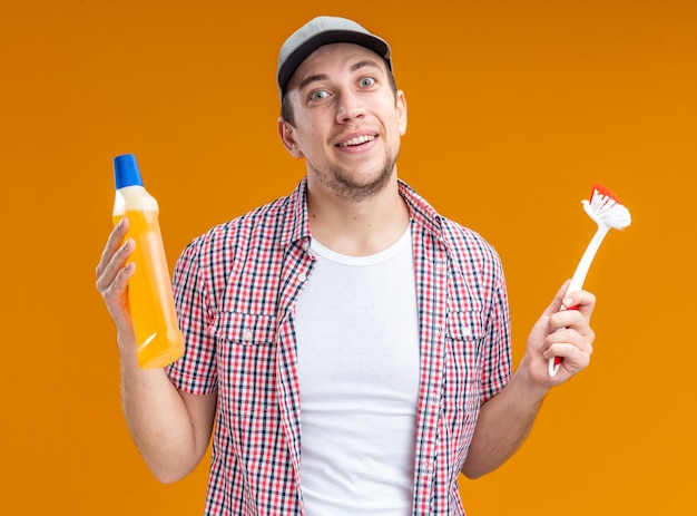 Sorridente giovane pulitore che indossa un cappuccio che tiene un agente di pulizia con una spazzola isolata su sfondo arancione