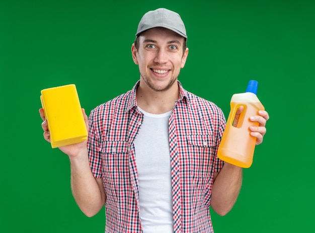 Sorridente giovane pulitore che indossa un cappuccio che tiene un agente di pulizia con una spugna isolata su sfondo verde