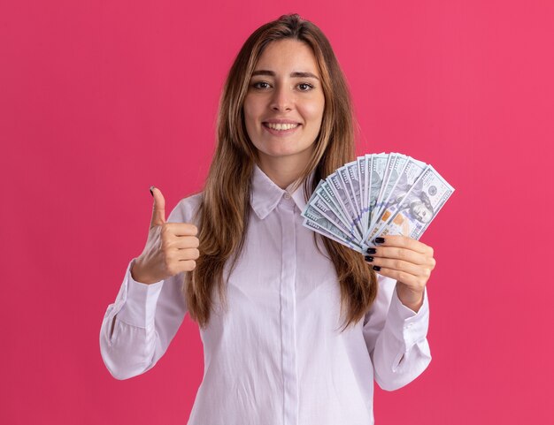 Sorridente giovane ragazza abbastanza caucasica thumbs up e tiene i soldi sul rosa