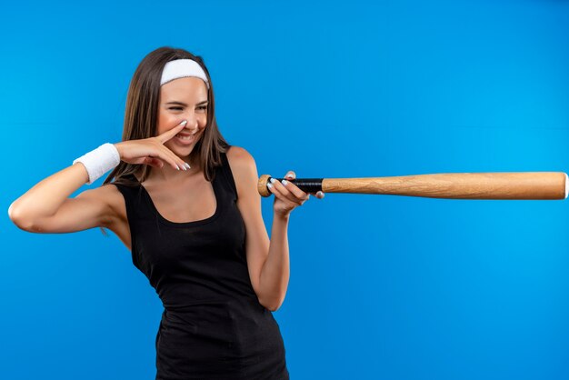 Sorridente giovane ragazza abbastanza sportiva che indossa la fascia e il braccialetto alla ricerca e che punta a lato con la mazza da baseball con il dito sul naso isolato sullo spazio blu