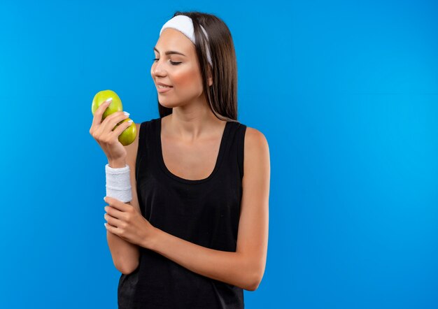 Sorridente giovane ragazza abbastanza sportiva che indossa la fascia e il braccialetto che tengono le mele guardandole e tenendo il braccio isolato sullo spazio blu