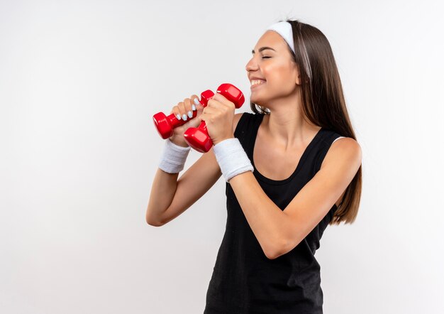 Sorridente giovane ragazza abbastanza sportiva che indossa la fascia e il braccialetto tenendo i manubri con gli occhi chiusi isolati su uno spazio bianco