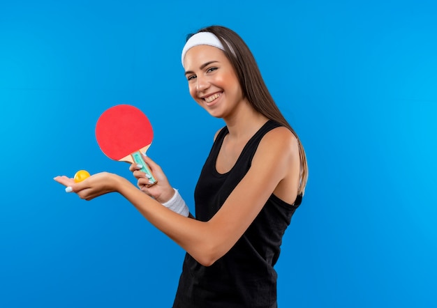 Sorridente giovane ragazza abbastanza sportiva indossando la fascia e il braccialetto che tiene le racchette da ping pong e la palla sullo spazio blu