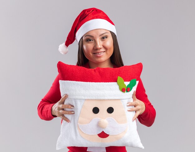 Sorridente giovane ragazza asiatica che indossa cappello di natale con maglione che tiene cuscino di natale isolato su sfondo bianco