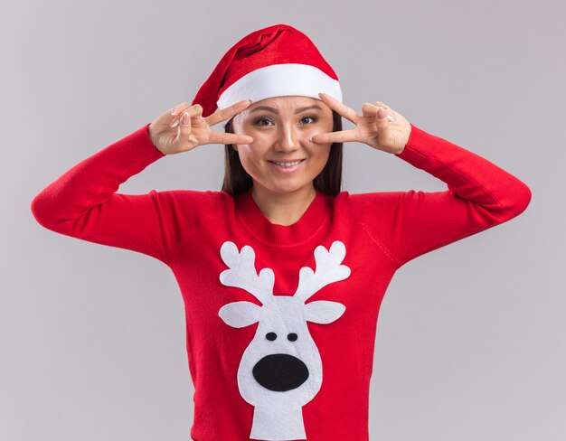 Sorridente giovane ragazza asiatica che indossa il cappello di Natale con il maglione che mostra il gesto di pace isolato su sfondo bianco