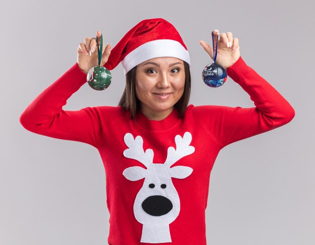 Sorridente giovane ragazza asiatica che indossa il cappello di Natale con il maglione che tiene le palle dell'albero di Natale isolato su sfondo bianco