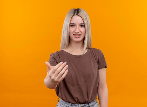 Sorridente giovane ragazza bionda con parentesi graffe dentali facendo venire qui gesto sullo spazio arancione isolato con spazio di copia