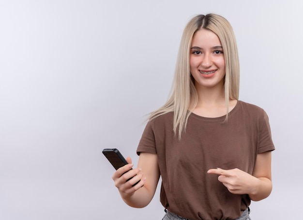 Sorridente giovane ragazza bionda in parentesi graffe dentale che tiene il telefono cellulare e indicandolo su uno spazio bianco isolato con spazio di copia