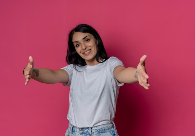 Sorridente giovane ragazza bruna caucasica allungando le mani fingendo di tenere qualcosa di isolato sulla parete rosa