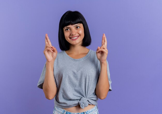 Sorridente giovane ragazza bruna caucasica incrocia le dita guardando il lato isolato su sfondo viola con spazio di copia