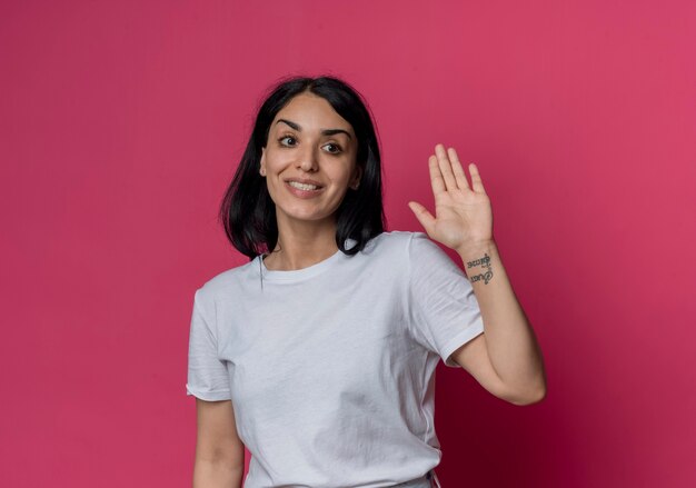 Sorridente giovane ragazza bruna caucasica solleva la mano isolata sulla parete rosa