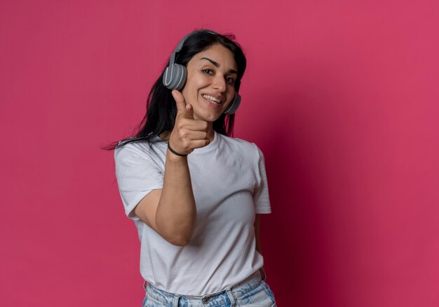 Sorridente giovane ragazza bruna caucasica sulle cuffie punti isolati sulla parete rosa