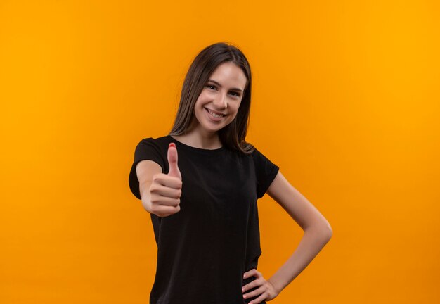 Sorridente giovane ragazza caucasica che indossa la maglietta nera con il pollice in alto mise la mano sul fianco su sfondo arancione isolato