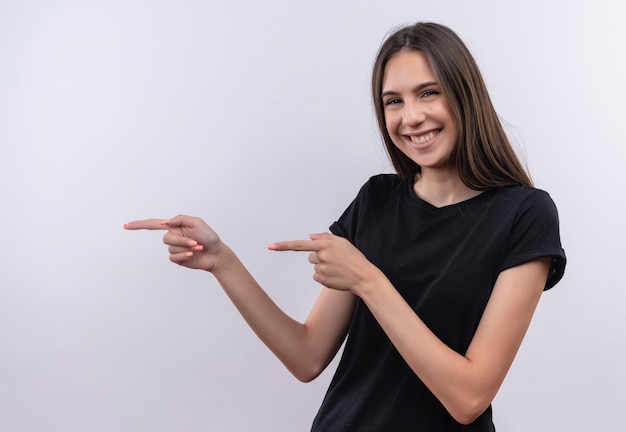 Sorridente giovane ragazza caucasica che indossa la maglietta nera punta le dita a lato su sfondo bianco isolato