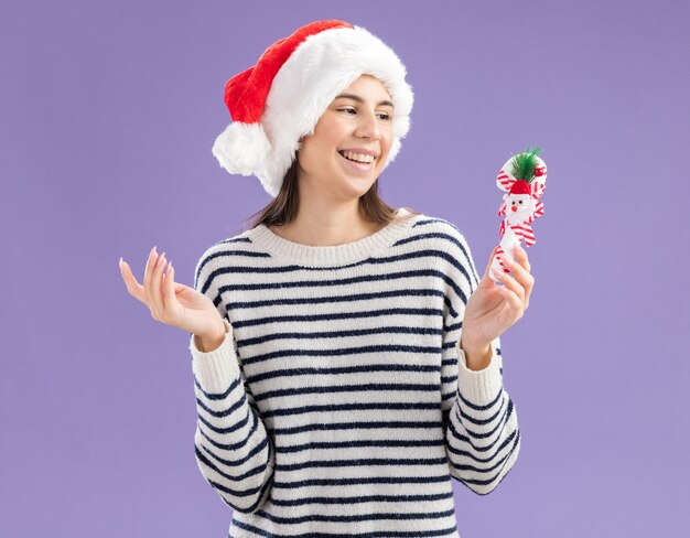 Sorridente giovane ragazza caucasica con cappello da Babbo Natale che tiene e guarda il bastoncino di zucchero isolato sul muro viola con spazio per le copie