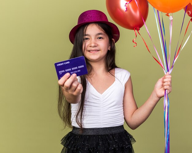 Sorridente giovane ragazza caucasica con cappello da festa viola in possesso di carta di credito e palloncini di elio isolati su parete verde oliva con spazio di copia