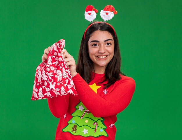 Sorridente giovane ragazza caucasica con fascia santa tenendo il sacchetto regalo di Natale isolato su sfondo verde con spazio di copia