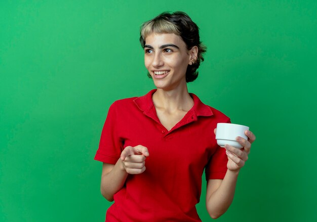 Sorridente giovane ragazza caucasica con pixie haircut holding cup guardando a lato e indicando la fotocamera isolata su sfondo verde con spazio di copia