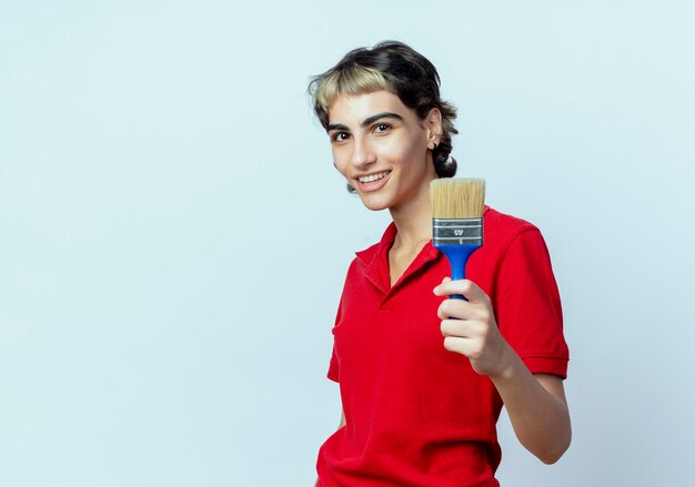 Sorridente giovane ragazza caucasica con taglio di capelli pixie tenendo il pennello isolato su sfondo bianco con spazio di copia