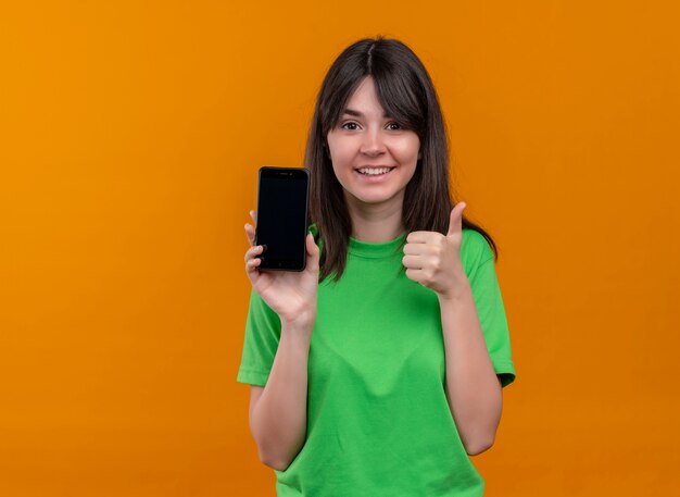 Sorridente giovane ragazza caucasica in camicia verde tiene il telefono e il pollice in alto su sfondo arancione isolato