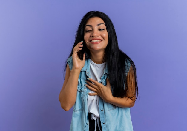 Sorridente giovane ragazza caucasica parla al telefono mette la mano sul petto isolato su sfondo viola con copia spazio