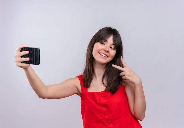 Sorridente giovane ragazza caucasica tenendo il telefono e facendo il gesto di pace isolato su sfondo bianco