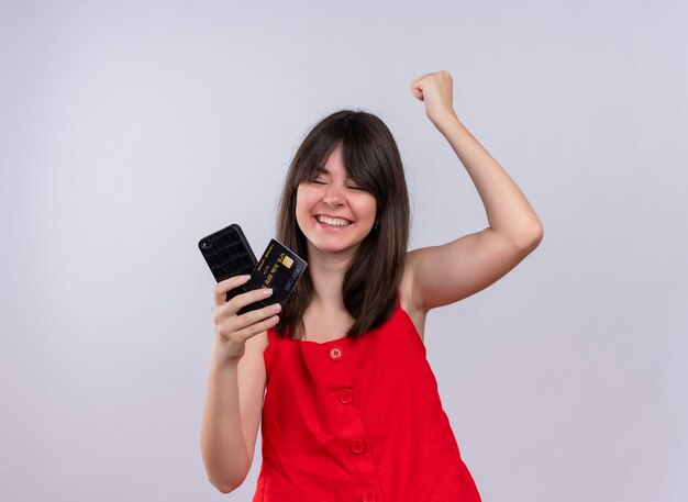 Sorridente giovane ragazza caucasica tenendo il telefono e la carta di credito insieme e alzando il pugno guardando il telefono su sfondo bianco isolato