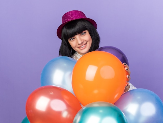 Sorridente giovane ragazza che indossa un cappello da festa in piedi dietro i palloncini che ne toccano uno isolato sul muro viola