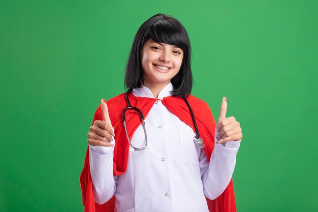 Sorridente giovane ragazza del supereroe che indossa uno stetoscopio con abito medico e mantello che mostra i pollici in su isolato sul verde