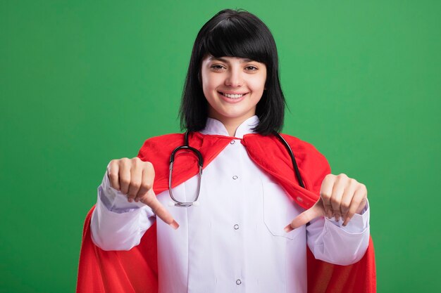 Sorridente giovane ragazza del supereroe che indossa uno stetoscopio con abito medico e mantello che mostra i pollici verso il basso isolato sulla parete verde