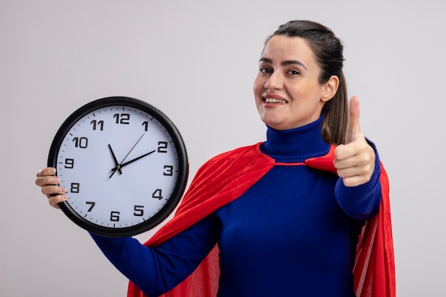 Sorridente giovane ragazza del supereroe tenendo l'orologio da parete che mostra il pollice in alto isolato su sfondo bianco