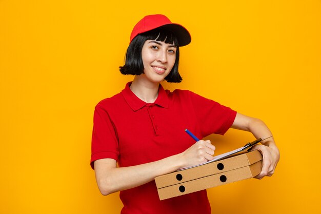 Sorridente giovane ragazza delle consegne caucasica che tiene in mano scatole per pizza e scrive negli appunti