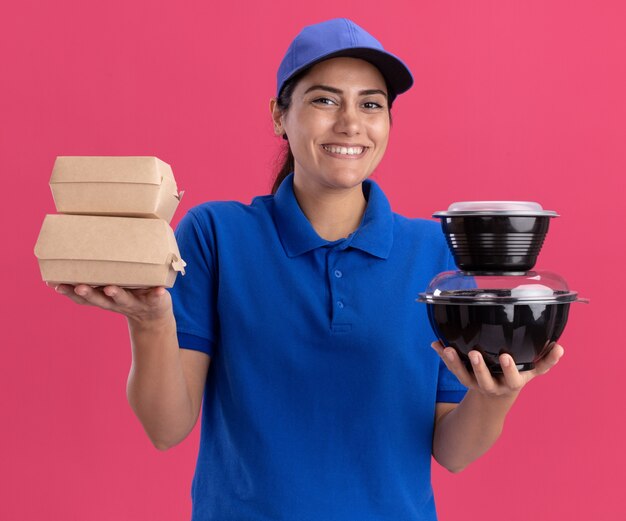 Sorridente giovane ragazza delle consegne che indossa l'uniforme con il cappuccio che tiene i contenitori per alimenti isolati sulla parete rosa