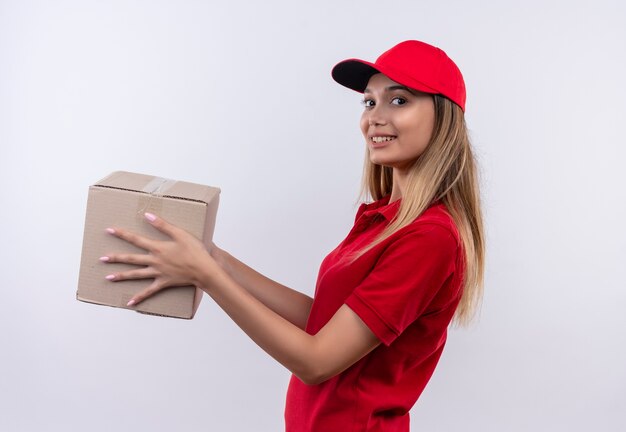 sorridente giovane ragazza delle consegne che indossa l'uniforme rossa e berretto tenendo fuori la scatola a lato isolato sul muro bianco