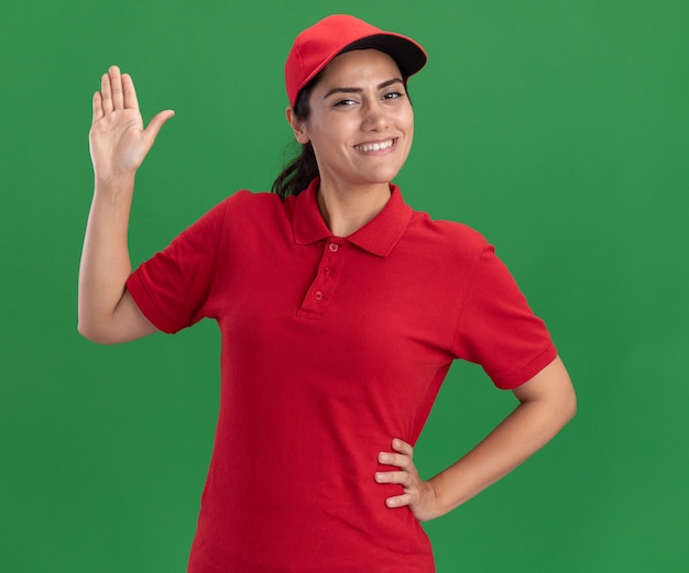 Sorridente giovane ragazza delle consegne indossando l'uniforme e cappuccio alzando la mano mettendo la mano sull'anca isolata sulla parete verde