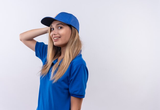 Sorridente giovane ragazza di consegna che indossa l'uniforme blu e cappuccio mettendo la mano sulla testa isolata su bianco