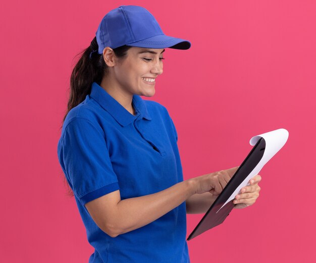 Sorridente giovane ragazza di consegna che indossa l'uniforme con cappuccio che tiene e guardando appunti isolato sulla parete rosa