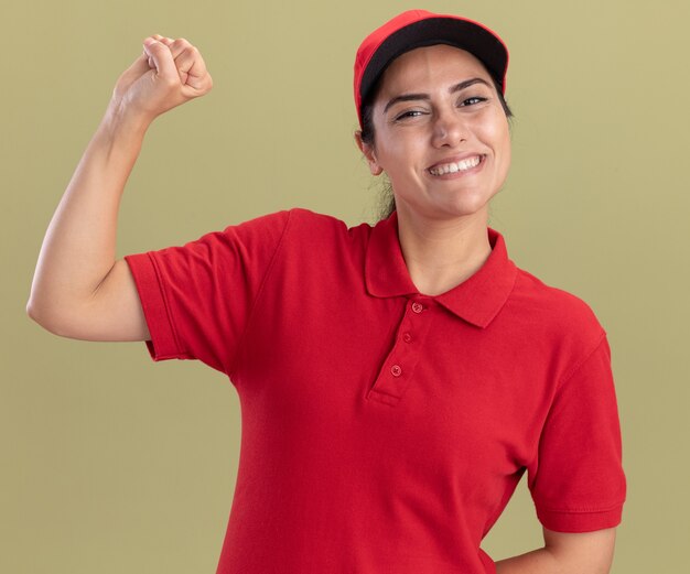 Sorridente giovane ragazza di consegna che indossa l'uniforme con cappuccio facendo forte gesto isolato sulla parete verde oliva