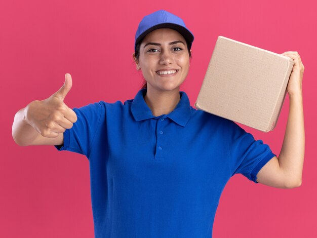 Sorridente giovane ragazza di consegna che indossa l'uniforme con cappuccio tenendo la scatola sulla spalla e mostrando il pollice in alto isolato sulla parete rosa