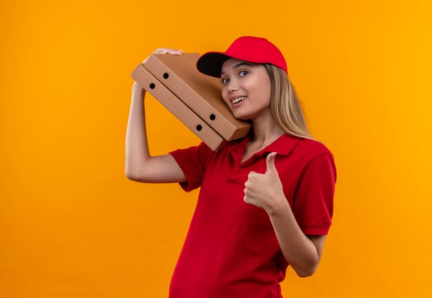 Sorridente giovane ragazza di consegna che indossa l'uniforme rossa e berretto che tiene la scatola della pizza sulla spalla il suo pollice in alto isolato sulla parete arancione