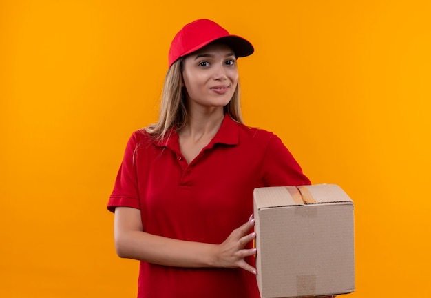 Sorridente giovane ragazza di consegna che indossa l'uniforme rossa e berretto che tiene scatola isolata sulla parete arancione