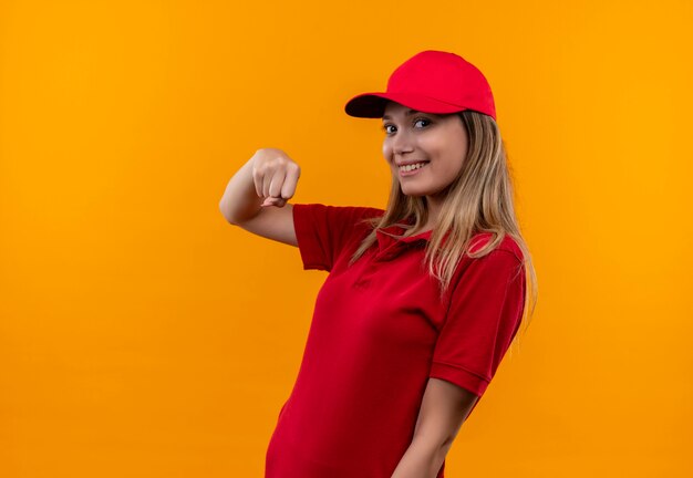 Sorridente giovane ragazza di consegna che indossa l'uniforme rossa e cappuccio che mostra il pugno isolato sulla parete arancione