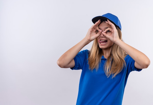 Sorridente giovane ragazza di consegna che indossa uniforme blu e cappuccio che mostra la lingua e guardando il gesto isolato su bianco