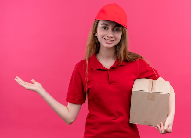 Sorridente giovane ragazza di consegna in rosso uniforme che tiene la scatola e mostrando la mano vuota sullo spazio rosa isolato