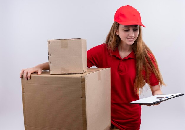 Sorridente giovane ragazza di consegna in rosso uniforme tenendo scatole e appunti e guardando appunti su spazio bianco isolato