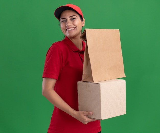 Sorridente giovane ragazza di consegna indossando l'uniforme e la scatola di contenimento del cappuccio con il pacchetto di cibo di carta isolato sulla parete verde