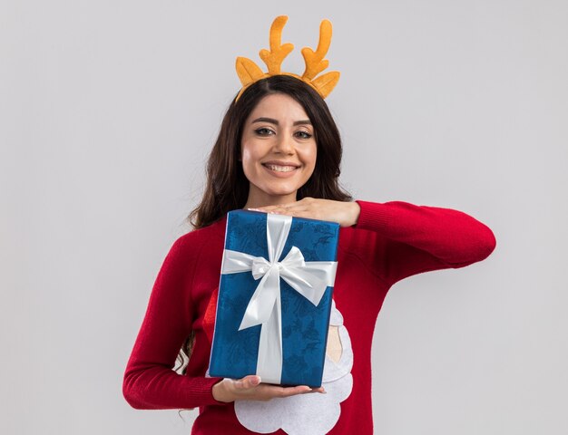 Sorridente giovane ragazza graziosa che indossa la fascia di corna di renna e maglione di Babbo Natale che tiene il pacchetto regalo di Natale alla ricerca