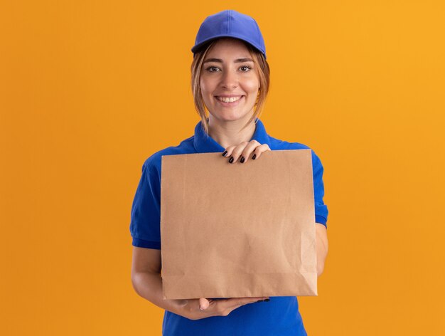 Sorridente giovane ragazza graziosa di consegna in uniforme che tiene il pacchetto di carta sull'arancio